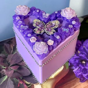 Könnte beinhalten: Eine herzförmige, lavendelfarbene Schmuckschatulle mit einem dekorativen Schmetterling und floralen Verzierungen. Die Schatulle ist mit einer silbernen Zierleiste versehen und verfügt über einen juwelenbesetzten Schmetterling als Mittelstück, umgeben von lila und weißen Rosen.