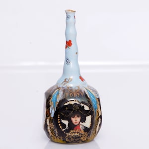 May include: A small, blue and white porcelain vase with a long, slender neck. The vase is decorated with gold accents and a painted image of a woman wearing a black hat and a red scarf.
