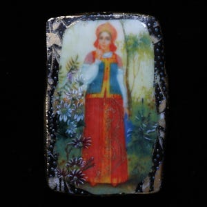 May include: A rectangular ceramic tile with a gold border and a painted image of a woman in a red dress and blue jacket standing in a forest. The woman is wearing a red headscarf and has a white collar. The background is a green forest with white flowers.