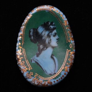 Op de afbeelding: Ovale porseleinen cameo met een gouden rand. De cameo toont een portret van een vrouw in een blauw-witte jurk tegen een groene achtergrond.