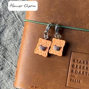 Könnte beinhalten: Zwei orangefarbene Planer-Anhänger in Toastform mit einem Bärengesicht, befestigt an einem silbernen Verschluss. Die Anhänger hängen an einem braunen Lederjournal. Der Text "Planner Charm" befindet sich in der oberen linken Ecke.