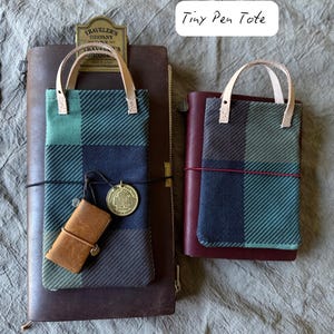 May include: Two pen totes with plaid patterns in shades of green and blue, featuring leather handles. One tote is paired with a brown leather notebook and a small leather pen holder. The text "Tiny Pen Tote" is visible.