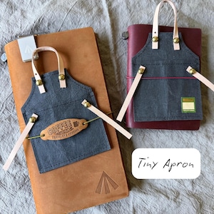 May include: Two leather notebooks with tiny gray aprons attached. The aprons have tan straps and brass rivets. One apron has a tag that reads "Coffee and Traveler's Notebook". The notebooks are brown and burgundy.