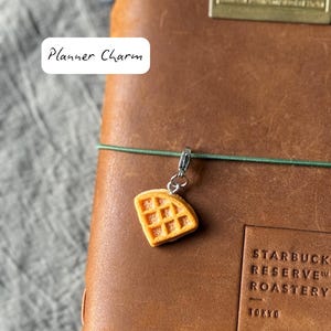 May include: A small, orange waffle charm with a silver clasp hangs from a green string on a brown leather notebook. The text "Planner Charm" is in a white box. The notebook has the words "Starbucks Reserve Roastery Tokyo" embossed on it.