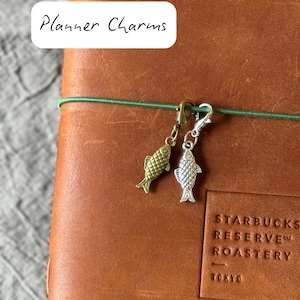 Puede incluir: Dos dijes de planificador con forma de pez, uno dorado y otro plateado, cuelgan de una banda elástica verde en un diario de cuero marrón. El texto "Planner Charms" está en un recuadro blanco. El diario tiene el logotipo de Starbucks Reserve Roastery Tokyo.