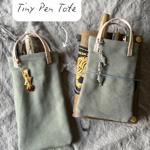 Puede incluir: Dos pequeños estuches para bolígrafos de color verde salvia con asas de color beige claro y abalorios decorativos. Los estuches tienen forma rectangular y están hechos de una tela texturizada. El texto "Tiny Pen Tote" está escrito en cursiva encima de los estuches.