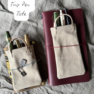 Può includere: Due piccole borse in tela beige per penne, con manici in pelle, una con un ciondolo a forma di chitarra. Una borsa è su un taccuino in pelle bordeaux, l'altra su un taccuino marrone. Le borse contengono penne e matite.