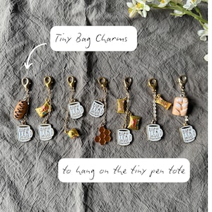 Puede incluir: Una colección de pequeños dijes para bolsos con cierres dorados. Los dijes incluyen alimentos en miniatura como un cruasán, caramelos y una taza de té con el texto "A cup of tea solves everything". El texto "Tiny Bag Charms" está escrito arriba.