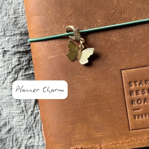 Puede incluir: Un dije de mariposa dorada sujeto a una banda elástica verde, apoyado sobre una agenda de cuero marrón. El texto "Planner Charm" está en un rectángulo blanco. La agenda tiene un borde cosido y texto en relieve.