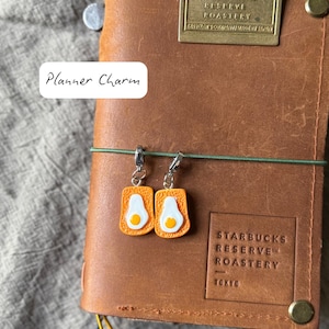 Könnte beinhalten: Ein braunes Lederjournal mit einem grünen Gummiband und einem Metallanhänger. Zwei kleine Anhänger in Form von Toast mit Spiegeleiern hängen an dem Journal. Der Text "Planner Charm" befindet sich auf einem weißen Etikett.