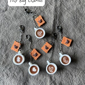 May include: Tiny bag charms featuring miniature coffee cups with latte art and small biscuit charms. The charms are attached to silver-colored chains and clasps. The text "Tiny Bag Charms" is in the upper left corner.