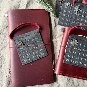 May include: A burgundy leather journal with a small, dark gray calendar tag attached by a red string. The tag has white numbers and a decorative charm. Several similar journals and tags are visible, along with a sprig of greenery.