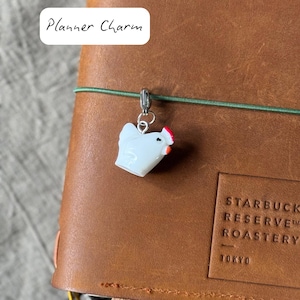 Può includere: Un ciondolo per planner a forma di pollo bianco con cresta rossa è appeso a un elastico verde su un diario in pelle marrone. Il testo "Planner Charm" è in un rettangolo bianco in alto a sinistra.