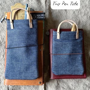 以下が含まれることがあります： デニム生地のTiny Pen Toteが2つ。革製のハンドルとアクセントが付いています。1つは茶色の革製ジャーナルに、もう1つはバーガンディ色の革製ジャーナルにセットされています。「Tiny Pen Tote」の文字が右上隅にあります。