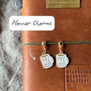 May include: Two gold-colored planner charms shaped like white mugs with the text "A cup of tea solves everything." The charms are attached to a brown leather notebook with the text "Planner Charms" in a white box.