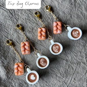 Może przedstawiać: Małe zawieszki do toreb z miniaturowymi filiżankami do kawy z latte art i pomarańczowymi kształtami plecionego chleba. Zawieszki są przymocowane do złotych łańcuszków z zapięciami na karabińczyk. Widoczny jest tekst "Tiny Bag Charms".