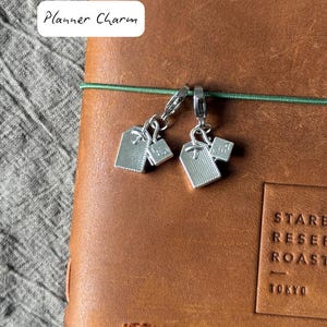 May include: Two silver-colored planner charms with a tag design are attached to a brown leather journal. The charms are hanging from a green elastic band. The text "Planner Charm" is in a white rectangle in the upper left corner.