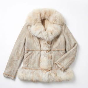 May include: A beige shearling coat with a large fur collar and trim. The coat features a button closure and decorative leather accents. The fur is a light cream color, and the coat appears to be made of soft, supple leather.