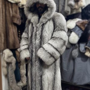 May include: A long, silver-grey fur coat with a large hood. The coat has a vertical striped pattern. Other fur coats are visible in the background. The coat is worn by a person.