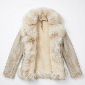 May include: A cream-colored shearling coat with a large fur collar and trim. The coat has a light brown suede exterior and a plush, off-white interior. The fur trim is a mix of cream and light brown.
