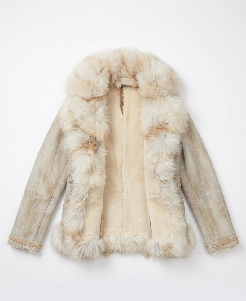 May include: A cream-colored shearling coat with a large fur collar and trim. The coat has a light brown suede exterior and a plush, off-white interior. The fur trim is a mix of cream and light brown.