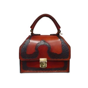 May include: A brown leather handbag with a top handle and a decorative lock. The bag has a unique design with black leather accents.