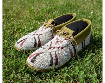 May include: A pair of beaded moccasins with a white base, adorned with red, black, and brown geometric patterns. The shoes have olive green suede collars and laces, and are set on a bed of green grass.
