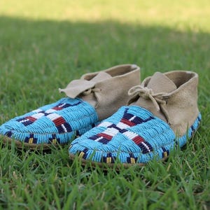 May include: A pair of beaded moccasin-style shoes with light tan suede uppers and turquoise blue beaded vamps. The vamps feature a geometric pattern in white, red, and navy blue. The shoes are set on a bed of green grass.