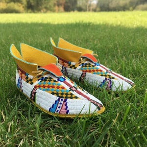 May include: A pair of beaded moccasins with a white base, featuring colorful geometric patterns in red, blue, yellow, and orange. The shoes have yellow collars and are set on a bed of green grass.