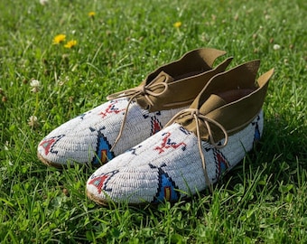 May include: A pair of white beaded moccasins with intricate patterns in red, blue, and black. The shoes have tan suede tops and laces, resting on a bed of green grass. The design features geometric shapes and tribal motifs.