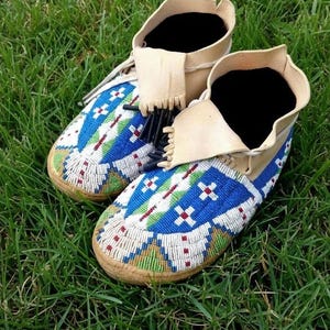 Puede incluir: Un par de mocasines hechos a mano con intrincados abalorios. El diseño presenta cuentas azules, blancas, rojas y verdes sobre una base de cuero beige. Los puños del tobillo son de cuero beige con flecos negros.