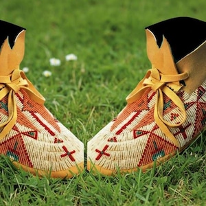 May include: A pair of beaded moccasins with a tan leather collar and laces. The moccasins are decorated with red, white, and orange geometric patterns. They are set on a bed of green grass.