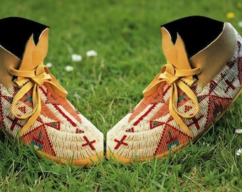 May include: A pair of beaded moccasins with a tan leather collar and laces. The moccasins are decorated with red, white, and orange geometric patterns. They are set on a bed of green grass.