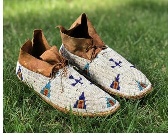 May include: A pair of handcrafted moccasins with a cream-colored beaded design. The moccasins feature brown leather tops and are decorated with blue, red, and yellow geometric patterns. They are displayed on a bed of green grass.