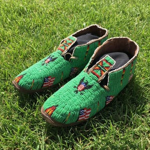 May include: A pair of green beaded moccasins with brown leather soles. The moccasins are decorated with American flags and eagle designs. The shoes are on a bed of green grass.
