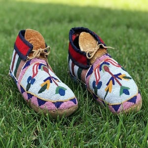 May include: A pair of beaded moccasins with a light blue background, featuring colorful floral patterns. The shoes have a red collar, brown leather lining, and are laced with tan string. The soles are a light brown color, and the shoes are set on green grass.