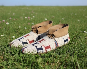 May include: A pair of beaded moccasins with a cream-colored base, featuring a pattern of red and navy blue animal figures. The moccasins have tan suede cuffs and laces. They are resting on a bed of green grass.