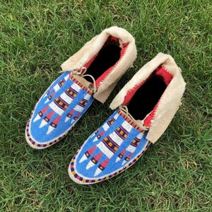 Puede incluir: Un par de mocasines con cuentas con un fondo azul y patrones geométricos en rojo, blanco y amarillo. Los zapatos tienen un ribete de piel color crema y forro rojo. Los mocasines se exhiben sobre hierba verde.