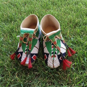 May include: A pair of beaded moccasins with a cream-colored interior and green, red, black, and white beadwork. The moccasins have a geometric design and red tassels. They are displayed on a bed of green grass.