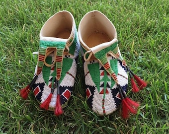 May include: A pair of beaded moccasins with a cream-colored interior and green, red, black, and white beadwork. The moccasins have a geometric design and red tassels. They are displayed on a bed of green grass.
