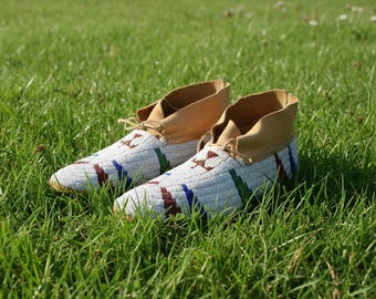 May include: A pair of beaded moccasins with tan leather tops and laces, resting on a bed of green grass. The white beaded uppers feature geometric patterns in red, blue, and green.