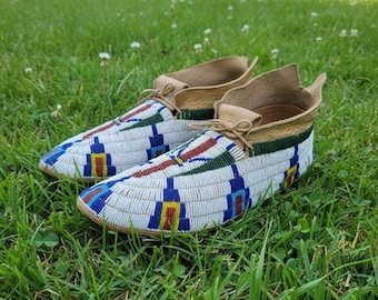 May include: A pair of beaded moccasins with a white base, adorned with blue, red, green, and yellow geometric patterns. The shoes have tan leather uppers and soles, and are tied with tan laces. The moccasins are resting on a bed of green grass.