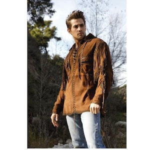 May include: Brown suede jacket with fringe detailing on the sleeves and chest. The jacket has a collared neckline and a lace-up closure. The model is wearing light blue jeans. The jacket is a western-inspired style.
