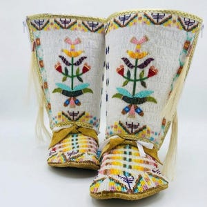 May include: A pair of tall boots featuring intricate beadwork in white, yellow, red, green, and blue. The design includes floral motifs, geometric patterns, and long fringe accents on the sides. The boots have tan leather details and white laces.