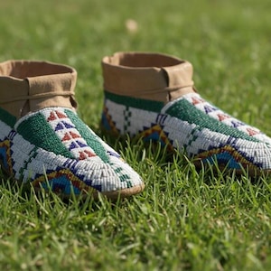May include: A pair of beaded moccasins with a tan leather collar and sole. The moccasins feature a white, green, blue, and red beaded design on the top and sides. The shoes are resting on a bed of green grass.