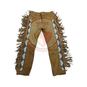Puede incluir: Pantalones de ante color canela con abalorios decorativos y flecos en los laterales. Los pantalones tienen cierre de botón y cremallera. El texto "NATIVE ZONE STORE" está impreso en los pantalones.