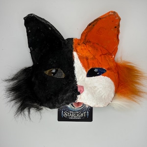 May include: A handmade cat mask, split vertically with black and orange fur. The left side is black, the right side is orange and white. The eyes are detailed with mesh and blue accents. The mask has a pink nose and a white muzzle. The text "Willow's Starlight Creations" is at the bottom.