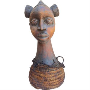 May include: A wooden sculpture of a woman's head with a basket base. The sculpture is carved in a dark brown wood and has a stylized face with large eyes and a wide mouth. The basket is woven from natural materials and is a dark brown color.