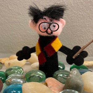 May include: A felt finger puppet of a wizard wearing round glasses, a black robe, and a red and yellow scarf. The puppet is holding a brown stick in its right hand. The puppet is surrounded by small, smooth stones.