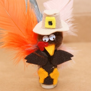 May include: A handcrafted Thanksgiving turkey decoration. The turkey is made of brown felt with a yellow beak and feet, black wings, and a red wattle. It wears a pilgrim hat and has orange and white feathers.